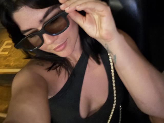 ScarlletZhen with dark hair, wearing black sunglasses and a black cut-out top, showing her tits, holding her glasses, and a gold chain, smiling. Tattoo on right wrist.