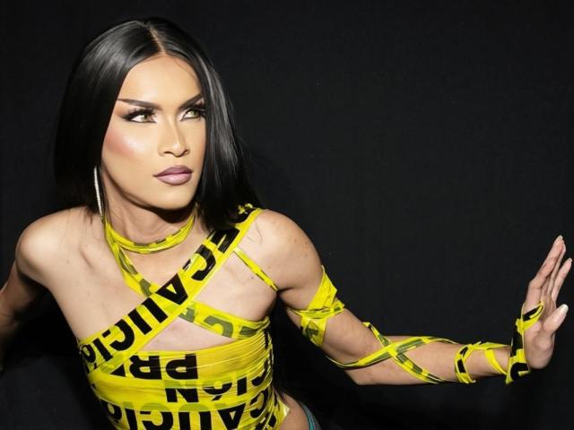 AgathaRodriguez has straight black hair, wearing a yellow "Caution" tape outfit with black text. Her makeup is bold, with dark eyebrows and pink lipstick. She's looking to the side with a serious expression. Her right hand is raised, palm facing forward. She has large silver hoop earrings. The "Caution" tape is wrapped around her torso, arms, and chest, partially covering her small breasts. The background is solid black, making her and the yellow tape stand out. Her skin is light brown. The image is sharp and well-lit.