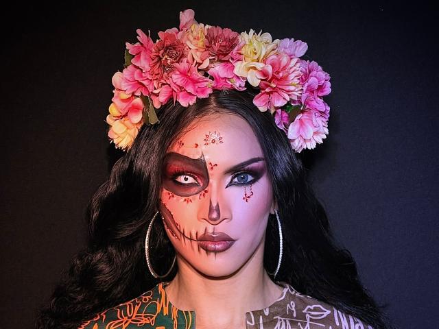 AgathaRodriguez has a dark, wavy hairdo and a flower crown with pink, orange, and yellow flowers. She has dramatic black eye makeup with red star patterns and black lipstick. Her right eye is partially covered by the makeup. She wears large silver hoop earrings and a brown, patterned top. The lighting is even, highlighting her makeup and floral crown. The composition is centered on her face, creating a strong visual focus.