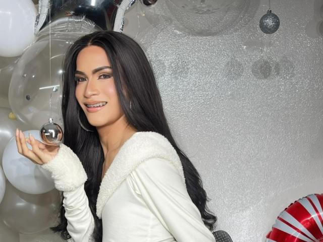 AgathaRodriguez with long black hair, white fluffy jacket, holding a silver ball, smiling, standing next to balloons, metallic decorations, and a red and white striped gift.