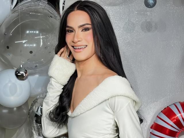 AgathaRodriguez with long black hair, smiling, wearing a white furry-sleeve top, showing off her tits, standing near balloons and a metallic sphere.