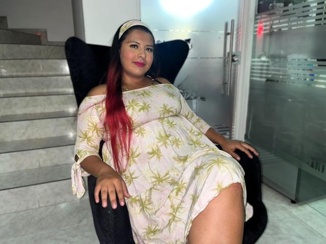 RileeyMoon, curvy with large tits, sitting on black chair, wearing off-shoulder, leaf-patterned dress, long red hair, white headband, pink lipstick, confident smile, marble stairs behind.