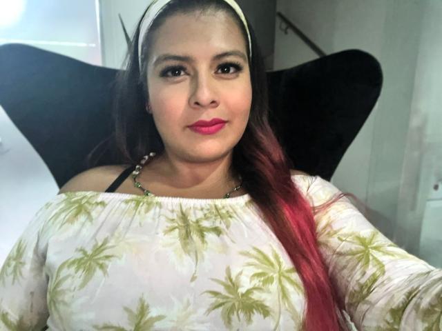 RileeyMoon with red lipstick, white headband, and green leaf pattern top, showing her tits, sitting in a black chair, looking at the camera, with long pink-dyed hair.