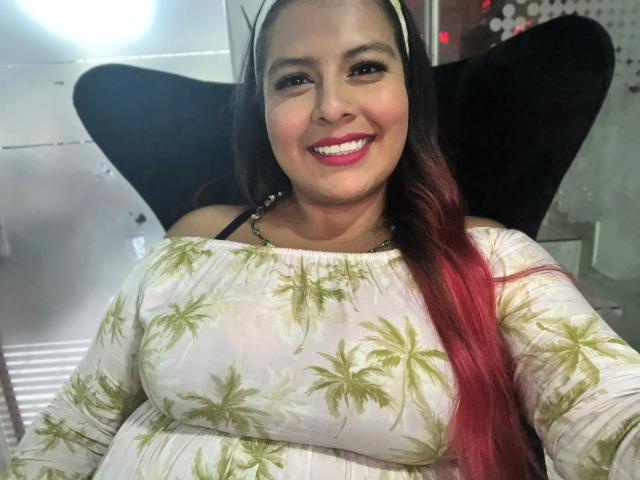 RileeyMoon with red-tipped long hair, smiling, wearing a white dress with green leaf patterns, showing her big tits, pink lipstick, black chair, and headband in a selfie.