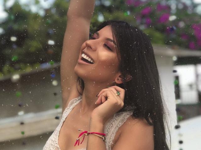 EmiliaWolf with dark hair, smiling, eyes closed, arm raised, lace top, red bracelet, green ring, white teeth, rain, bokeh, purple flowers in background.