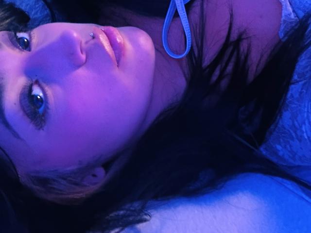 KatianPierce with purple-blue lit face, black hair, nose piercing, and blue eyes, lying down, wearing white lace top, looking up. Her tits are partially visible, arms raised.