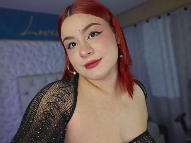 CassandraSexy has red hair, black lace top, small breasts, and a cute face with winged eyeliner, smiling slightly, wearing hoop earrings, in a softly lit room.