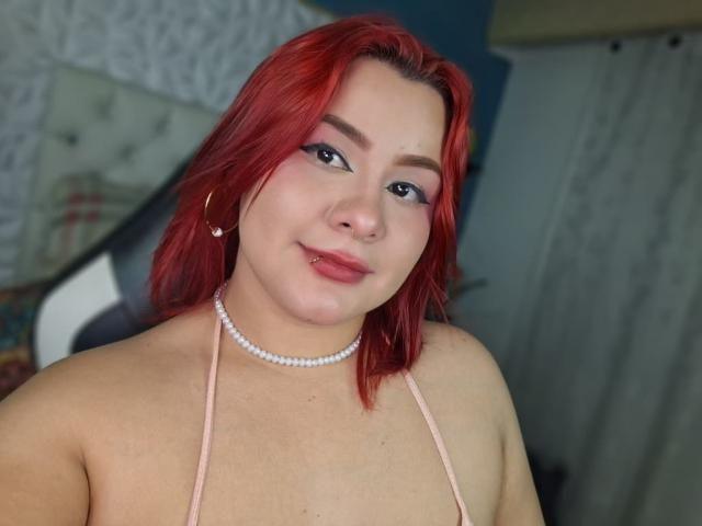 CassandraSexy with red hair, pink halter top, pearl necklace, hoop earrings, and blue eyeshadow, smiling at the camera, showing her tits and a nose ring.