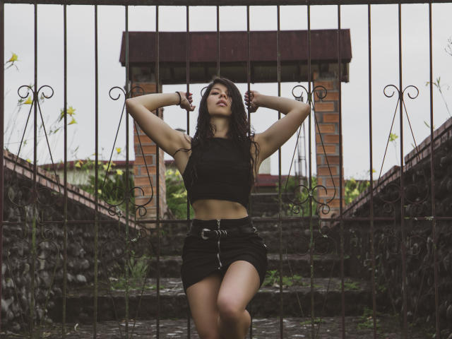 EmiliaWolf with arms raised, wearing a black crop top and mini skirt, standing in front of a metal gate, looking confident with a slight smirk, her tits visible.