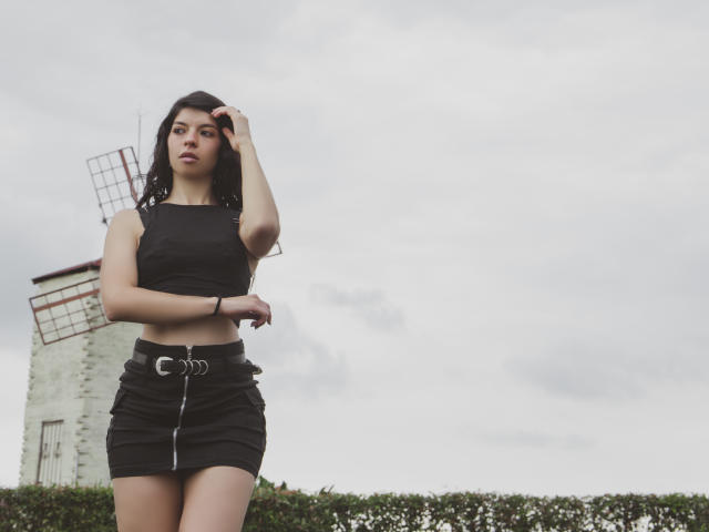 EmiliaWolf with long dark hair, wearing a black crop top and high-waisted black skirt with a belt, standing outside, hand in hair, looking down with a serious expression.