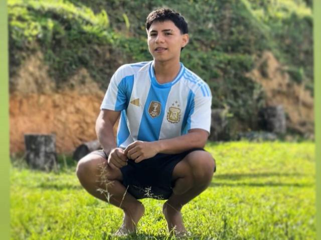 RichiiHott is squatting on grass, wearing a blue and white soccer jersey with a logo, black shorts, and no shoes, with short curly black hair and a slight smile.