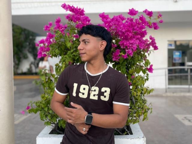 RichiiHott with dark short hair, brown t-shirt with "1923" in white, black watch, and necklace, standing in front of bright pink bougainvillea flowers, looking to the side.