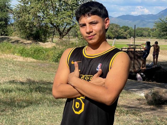 RichiiHott, muscular, tan-skinned, short black hair, sleeveless black sports jersey with yellow trim, standing with arms crossed, thumbs up, confident smile, outdoors, sunny, mountains in background.