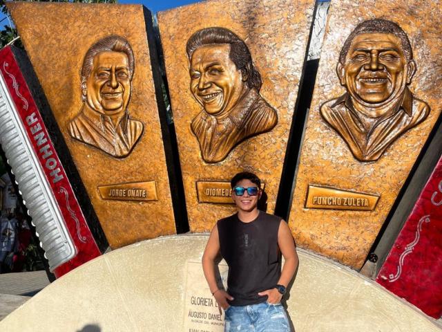 A photograph of RichiiHott standing in front of three wooden sculptures. RichiiHott, wearing a black sleeveless shirt and blue sunglasses, holds a sign. The sculptures are detailed carvings of Juanes, Simón Bolívar, and Pedro Pérez, each with a nameplate. The wood has a rough texture, and the carvings are in profile with smiling expressions. RichiiHott stands in a curved, beige stone structure. The wood is orange-brown with red accents on the sides.