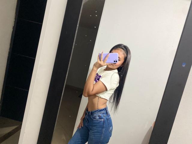 NatasshaAx in a mirror selfie, wearing a white crop top, blue high-waisted jeans, and a purple bracelet, with long black hair, showing her slim, toned, and flat-assed body.