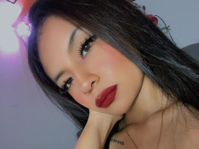 NatasshaAx has dark, straight hair, red lipstick, and a nose piercing. She's touching her chin with a hand, wearing long lashes, and has a tattoo near her collarbone.