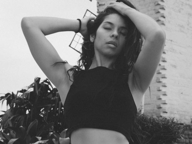 EmiliaWolf with wet hair, arms raised, eyes closed, wearing a black crop top, showing her small tits and toned midriff, standing near a plant, in a black-and-white photo.