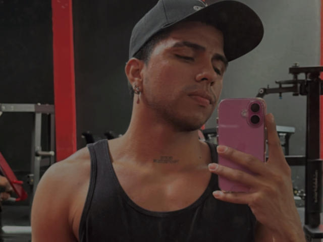 SebasAllen with a chiseled, muscular chest, black tank top, and cap, holding a pink phone, taking a selfie, showing a tattoo under his chin, in a gym.