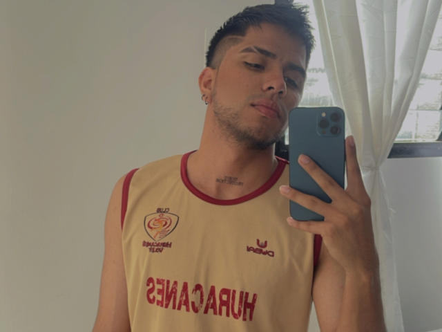 SebasAllen with short brown hair, light skin, and a beard, wearing a yellow sports tank with red trim, taking a selfie with a blue phone, pouty expression.