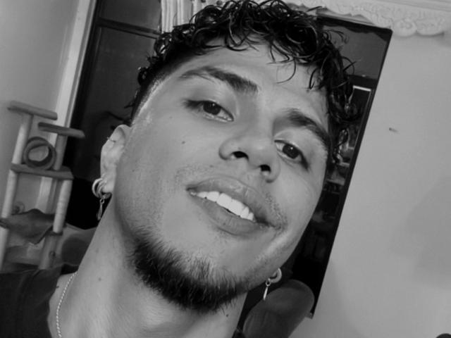 SebasAllen has curly black hair, a short beard, and a goatee. He's wearing a ring in his left ear and a necklace. His lips are slightly parted.