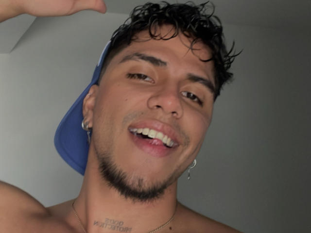 SebasAllen with a blue cap, black curly hair, nose ring, and small goatee, smiling with tongue out, showing a tattoo on his neck. Shirtless, gray background.