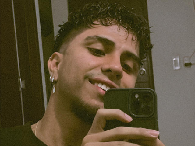 SebasAllen with short curly hair, ear gauges, and a black shirt, holding a phone, smiling, showing teeth, and wearing a necklace. His hand is near the phone's camera.