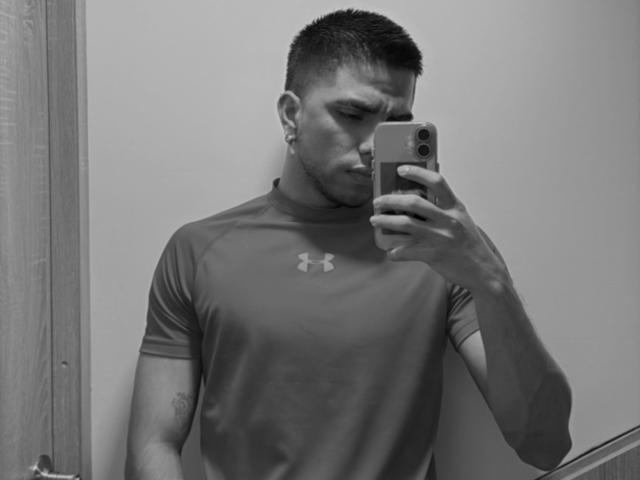 SebasAllen with short dark hair, Under Armour shirt, ear gauges, holding phone, taking mirror selfie, looking serious, tattoo on right arm, wood door on left. Black and white.