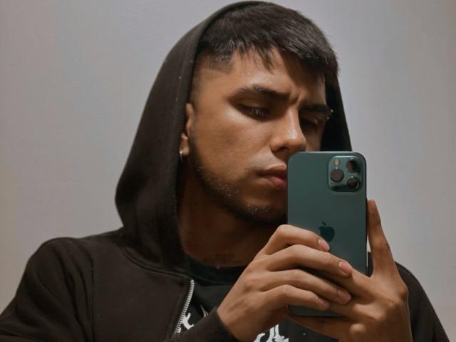 SebasAllen with short black hair, wearing a black hoodie, looking at a green iPhone with a serious expression, holding the phone in both hands, showing a small tattoo on neck.