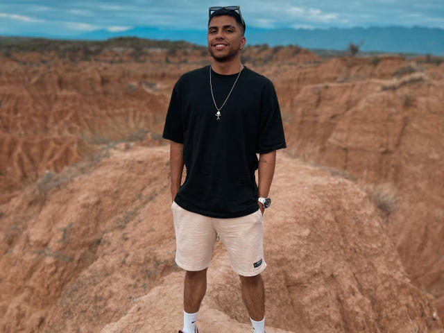 SebasAllen stands on a rocky outcrop, wearing a black T-shirt, beige shorts, and white socks. He has dark hair, a beard, and wears sunglasses on his head. A silver necklace with a star-shaped pendant hangs around his neck. His right hand is in his pocket. The rocky landscape in the background is rich in earthy reds and browns, with a cloudy blue sky overhead. The image has a natural, candid style.