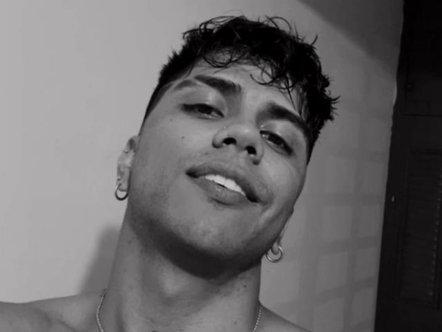 Black and white close-up of SebasAllen. SebasAllen has short, wet-looking dark hair and a slightly tilted head. He's wearing small hoop earrings in both ears and a thin chain necklace. His expression is relaxed, with a subtle smile and slightly parted lips. His right cheek is slightly puffed out. The lighting creates subtle shadows on his face and the wall behind him. The image has a casual, slightly rough texture. SebasAllen's skin tone is light to medium. The photograph focuses on his upper torso and face.