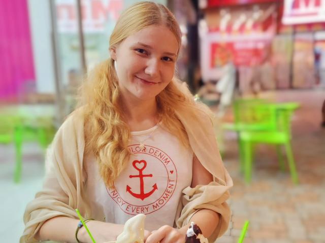 VanessaNorris with blonde pigtails, smiling, wearing a white shirt with a red anchor and heart, beige cardigan, holding a white napkin, sitting at a green chair.