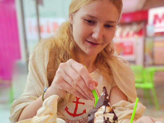 VanessaNorris with blonde hair, holding a dessert with chocolate and green straws, wearing a beige shirt with a red anchor, smiling, and looking down. Boobs slightly visible.