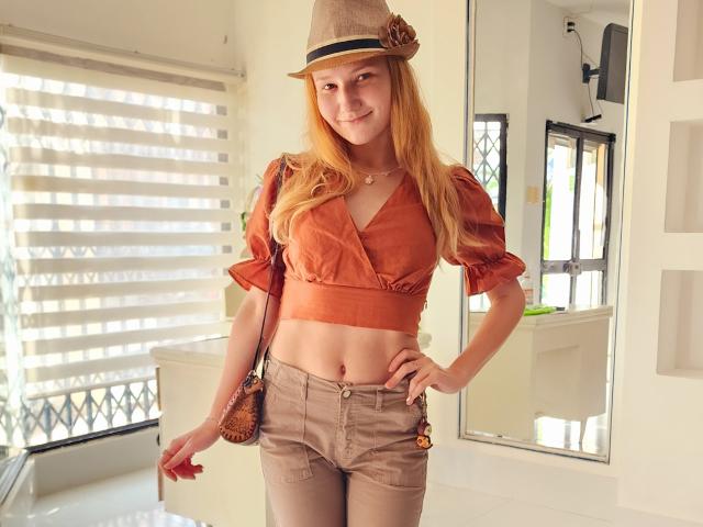 VanessaNorris with long red hair, wearing a brown hat, rust-colored crop top, and beige shorts, holding a brown purse, standing in a sunlit room, smiling.