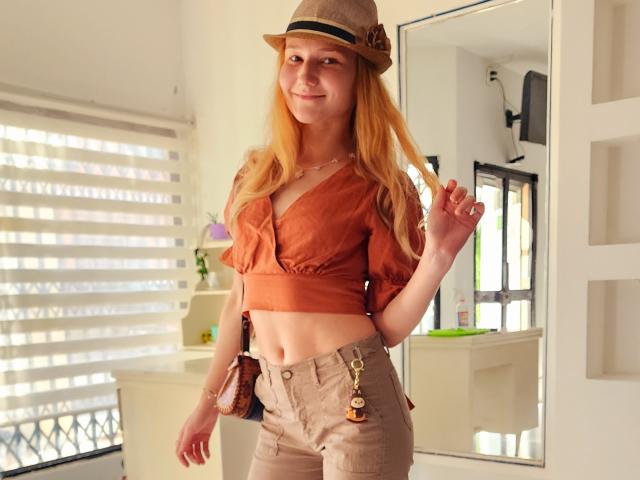 VanessaNorris with long blonde hair, brown hat, orange crop top, tan high-waisted pants, and a brown belt with a keychain, standing in a bright room.