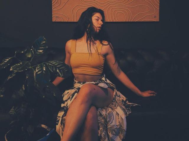 EmiliaWolf with long black hair, yellow crop top, and floral skirt, sitting on black couch, legs crossed, dark room, potted plant on left, orange wall art behind.