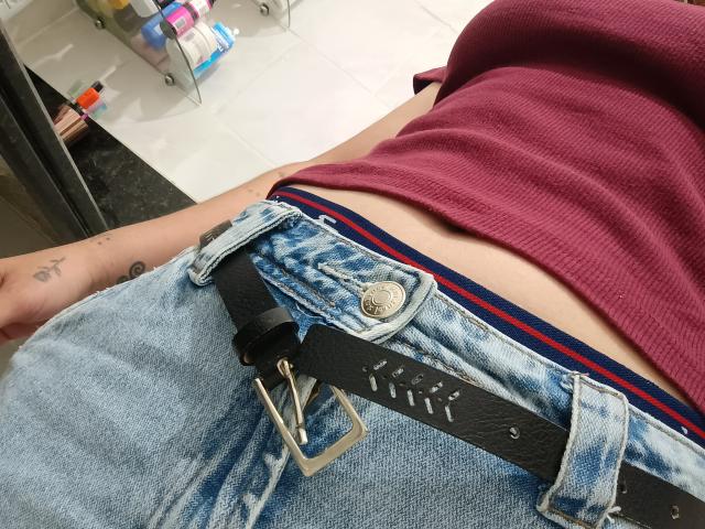 Le ventre apparent de RosyG, visible sous un haut rouge à rayures verticales, un jean en denim bleu avec une ceinture en cuir noir, révélant un tatouage sur son poignet gauche, et sa culotte noire..