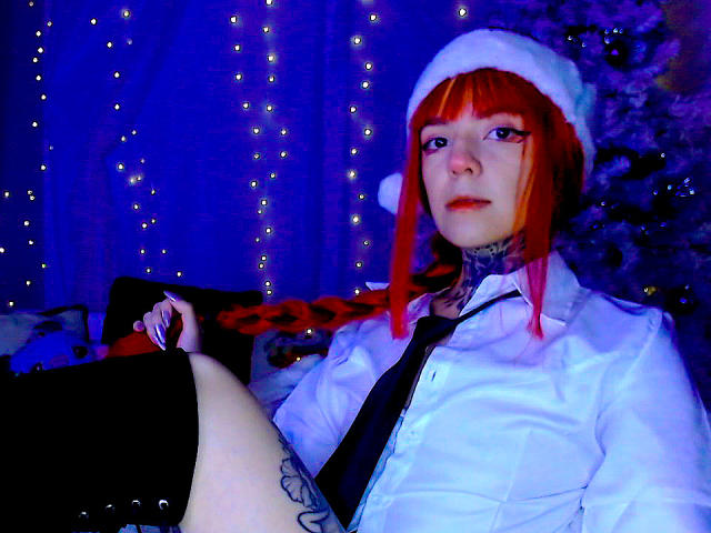 LannieGonzales with red hair in a white hat, white shirt, black tie, and black skirt, showing tattoos, sitting with legs apart, blue and purple lighting, looking at camera.
