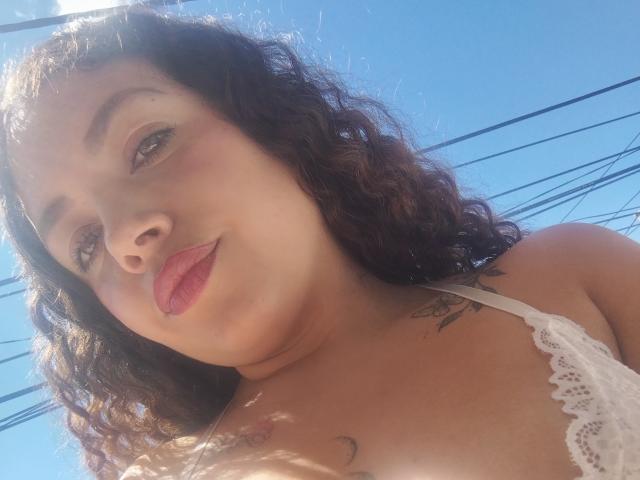 RubbyStrongX has curly brown hair, light brown skin, and pink lipstick. She's wearing a white lace top, showing small tits and a tattoo on her right chest.