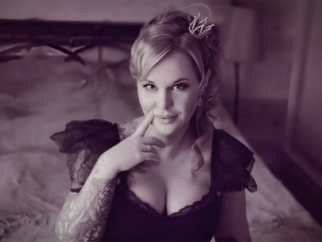 MissTwinkleX, a blonde woman with a crown in her hair, sits in a bed with a black iron headboard. She has tattoos on her right arm and is wearing a black, lace-trimmed dress with puffy sleeves. She has a slight smile, with her right index finger touching her chin. The image is in grayscale, emphasizing her light skin and dark dress. She has a nose ring and is looking directly at the camera. Her hair is styled in loose curls.