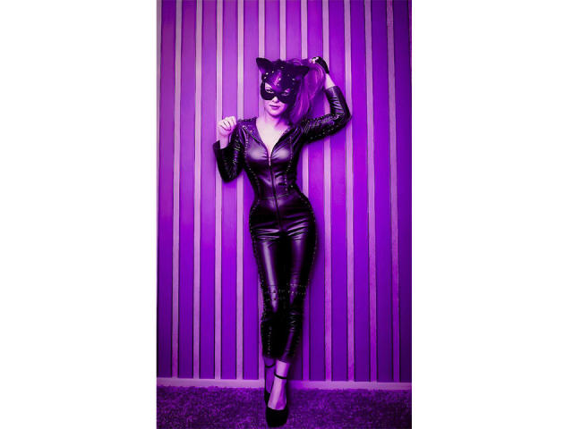 MissTwinkleX stands against a purple-striped wall, wearing a black leather cat mask and a tight, black leather catsuit. She has one hand up, gripping the mask, and the other rests on her hip. She's in black high heels, with purple lighting casting a neon glow. Her pink hair is tied back. The image is sharp, with a vibrant, purple-tinted color scheme. Her confident pose and sharp, shiny outfit dominate the composition.