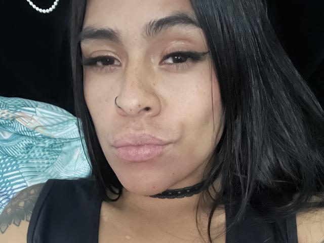 StellaBeckker with dark straight hair, nose ring, black choker, and winged eyeliner, pouting her lips, showing freckles on her nose and a tattoo on her left shoulder.