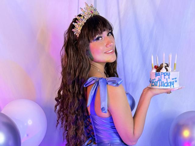HanaJoud with long wavy brown hair, wearing a glittery crown and blue satin dress, holding a birthday cake with candles, smiling at the camera, showing her ass.