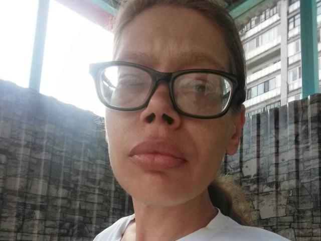 Lorraine Rux tiene una expresión seria, guiña un ojo, lleva gafas de color negro, cabello castaño claro recogido en una coleta y labios rosados. Lleva una camisa blanca..