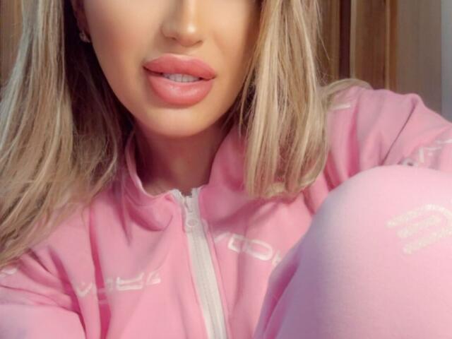 NicolSNaya's face with pink lips, blonde hair, wearing a pink zip-up hoodie, and pink knee-high socks, showing just her mouth, chest, and knee in a selfie.