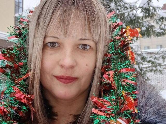 AngelaRoxyx mit blonden Haaren, die eine grüne und rote Girlande trägt, hat helle Haut, hellbraune Augen und roten Lippenstift und blickt direkt in die Kamera. Schnee ist im Hintergrund sichtbar..