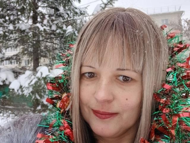 AngelaRoxyx with blonde hair, red and green tinsel around her head, wearing red lipstick, snow on her forehead, and a grey jacket, smiling softly. Snowy background.