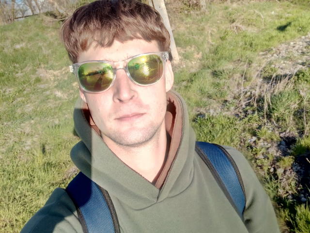 Luko6 9 has short brown hair and wears reflective green sunglasses. He's in a green hoodie with a brown inner lining, and a blue backpack. Sunlight highlights his face and glasses, creating a shiny reflection. The green grass in the background is slightly blurred. The image is a close-up selfie, focusing on Luko6 9's upper body and face.