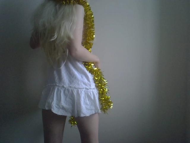 DitaVira's ass is covered by a white, short, sleeveless dress, holding a gold tinsel garland with long, blonde hair falling down her back.