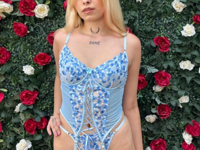 Lanna Hamilton in een blauwe kanten lingerie set met bloemmotieven, die haar borsten en schaamstreek laat zien, staand voor rode en witte rozen. Ze heeft blond haar en een maanvormige tatoeage..