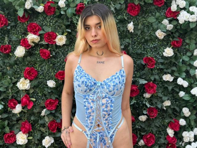 LannaHamilton with blonde hair, wearing a blue floral lace-up lingerie set, showing small tits, wearing a crescent moon necklace, standing in front of red and white roses.
